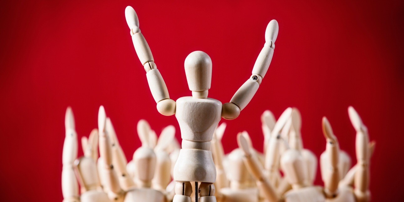 On a red background a wooden artist's mannequin stands with its arms raised addressing a crowd of other manequins.