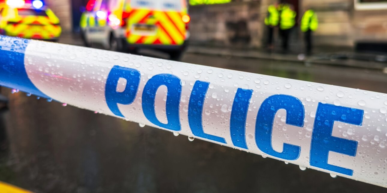 Blue and white striped tape with the word police written on it. In the background are several out-of-focus police officers wearing yellow hi-vis and two police vehicles