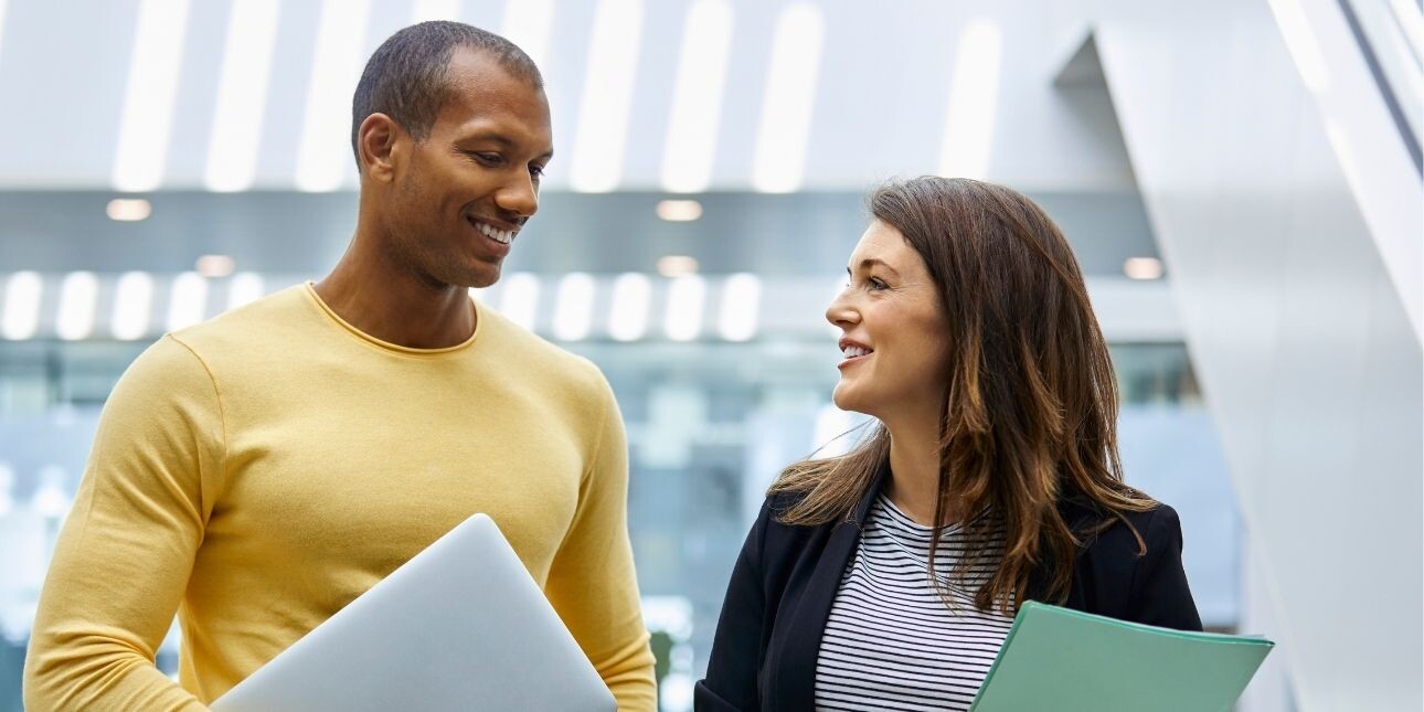 Two smiling colleagues - a Black man wearing a yellow jumper and a white woman wearing a dark jacket - chat in a light filled atrium. Both hold folders.
