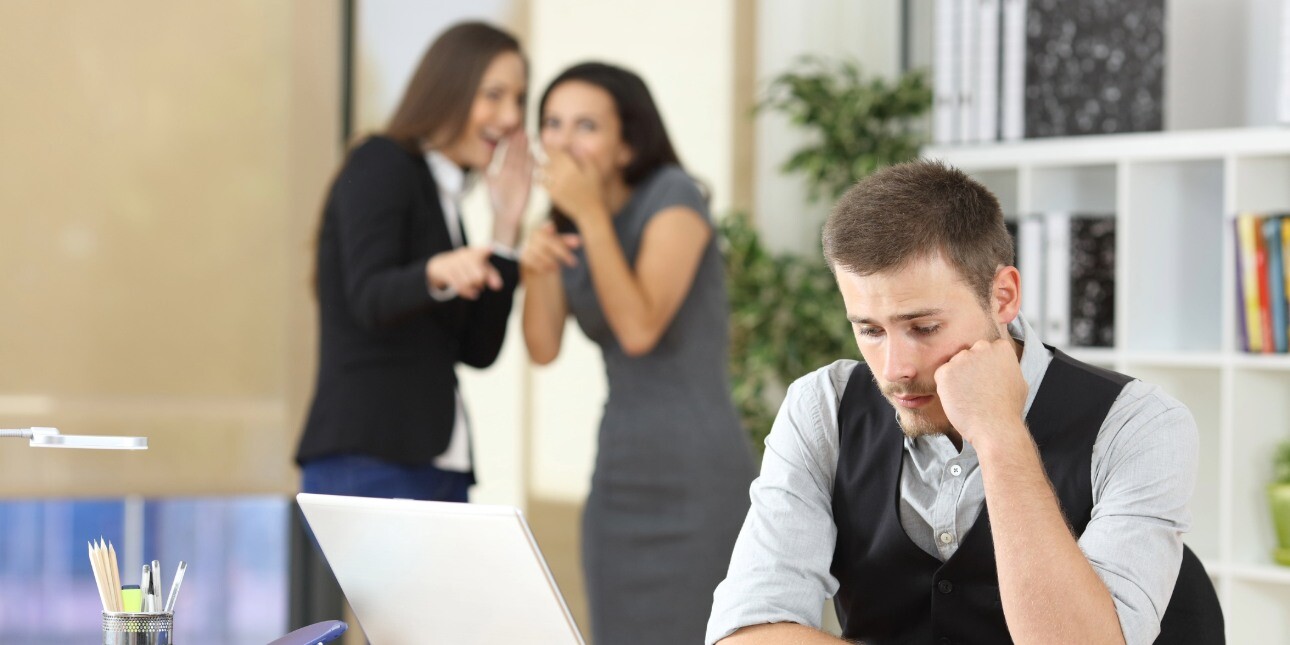 A white man rests his cheek on his hand as he stares dispondantly at his office desk while two women in the background whisper and laugh to each other.