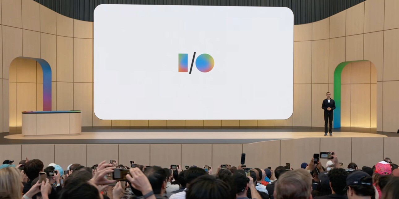 A screenshot from the Google I/O launch event with a shot across the backs of the audience towards a beige stage where a man stands to the right of a screen displaying the I/O logo.