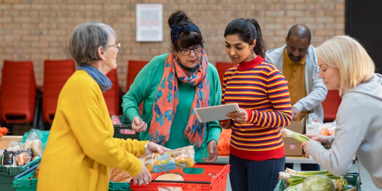 Five food bank volunteers of different ages, genders and ethnicities load food into boxes around tables.