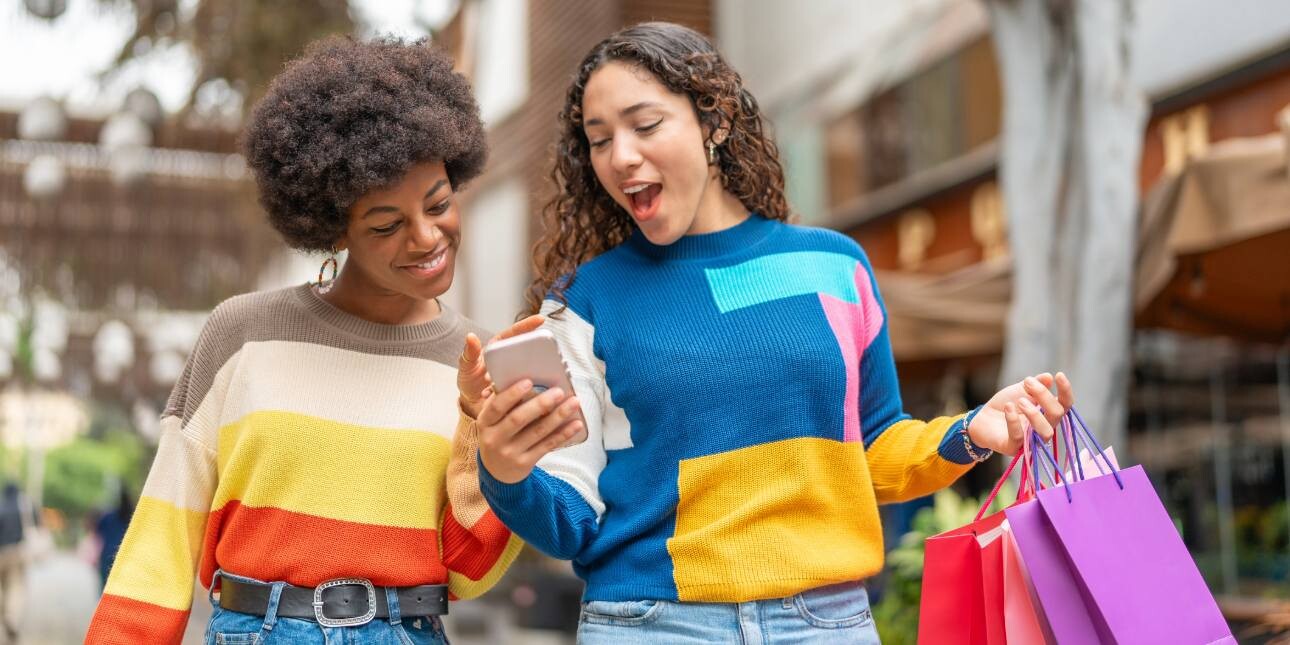 Two young women in brightly coloured jumpers smile while looking at the a mobile phone that one of them is holding.