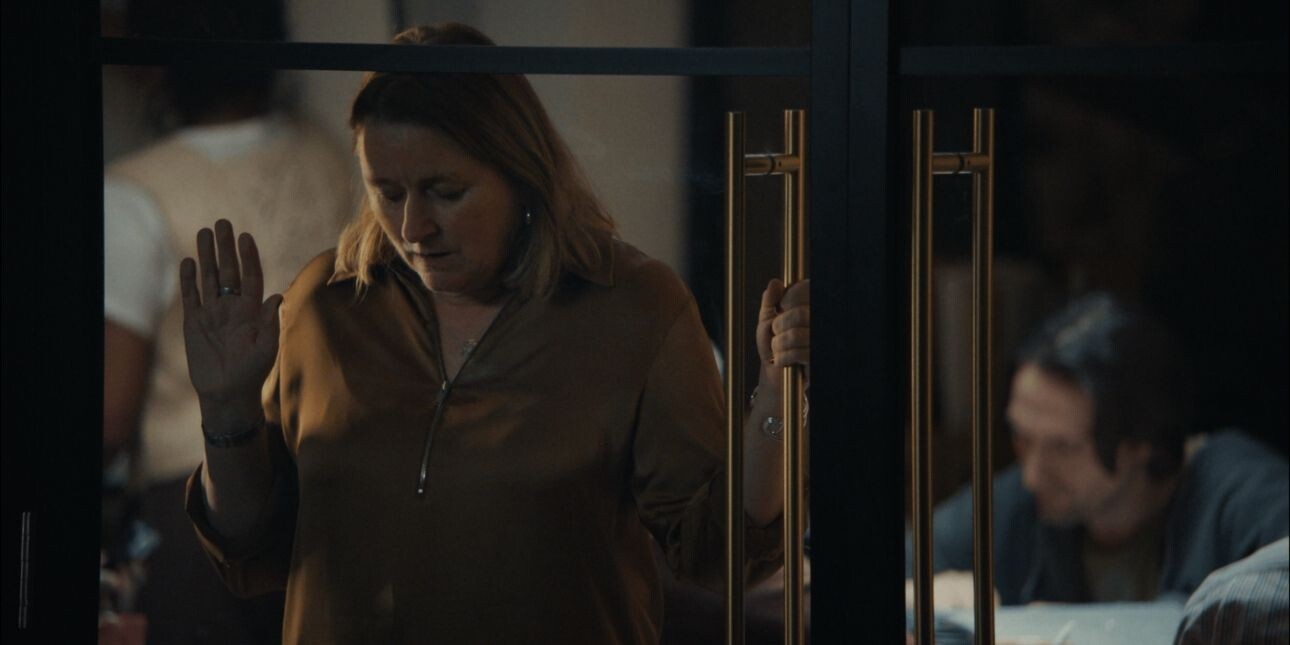 A white woman with blond hair rests her head and a raised palm on the inside of glass doors with gold handles. Two other people are stood and sat in the background.