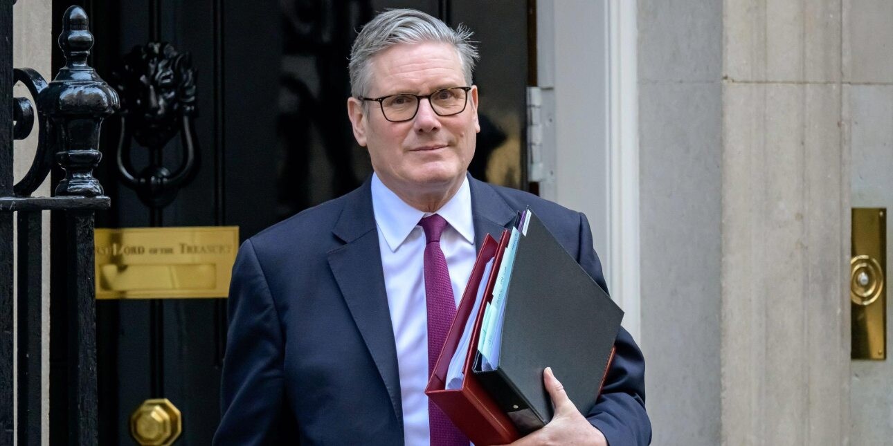British prime minister Keir Starmer wearing a grey suit and holding a red folder. Behind him is the black door to 10 Downing Street.