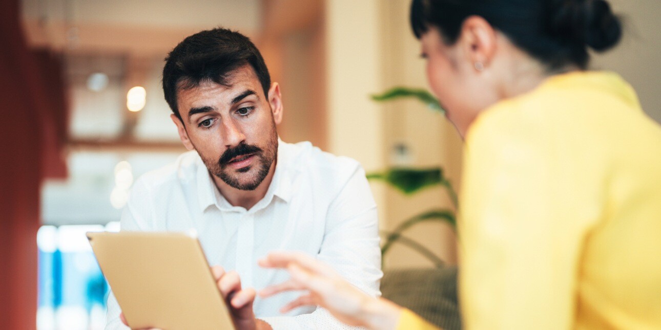 Two colleagues - a white man with dark hair and moustache - holds an iPad while in conversation with a female colleague with dark hair whose face is obscured.