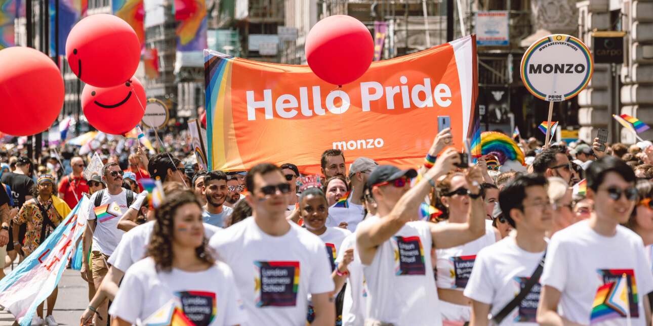 A group of people on a Pride march wearing white t-shirts. In the background an orange banner reads Hello Pride