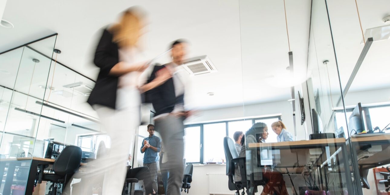 The blurred image of two colleagues walking through an office. Others are sat at desks in the background.