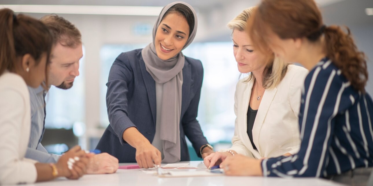 A woman wearing a grey hijab and blue outfit is stood over paperwork on a table while speaking to four male and female colleagues.