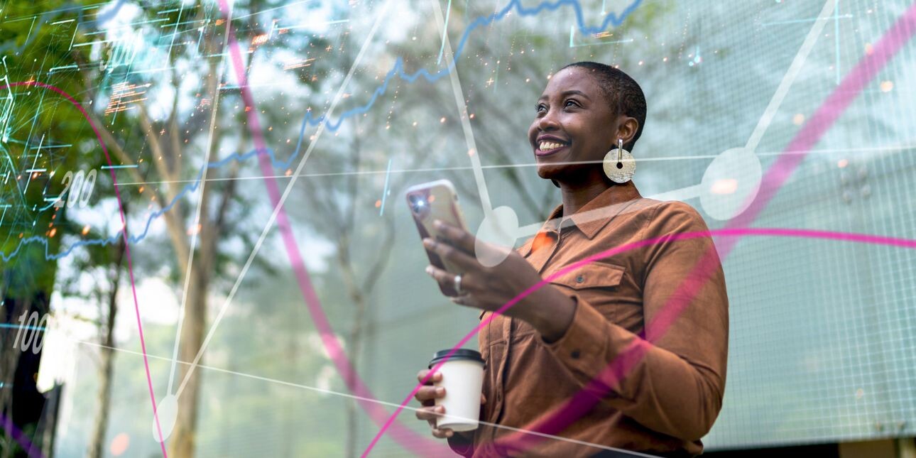 A smiling Black woman holds a phone in one hand and a coffee cup in the other, while stood beneath trees.