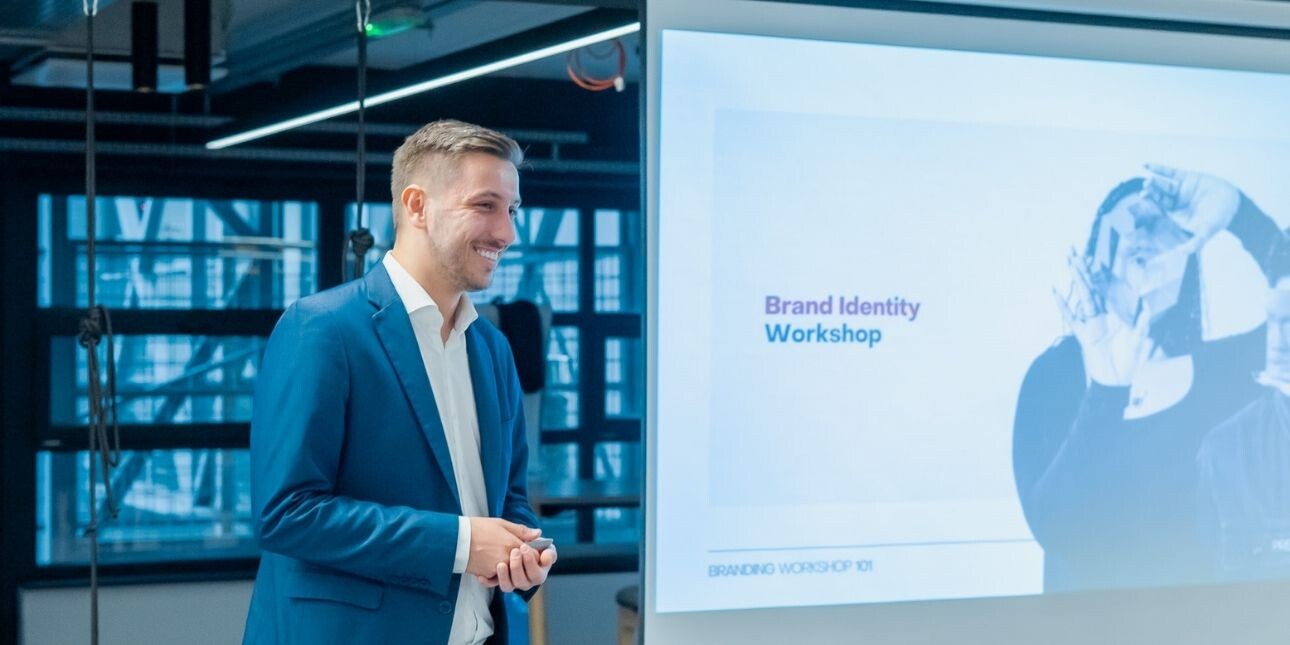 A colour portrait of a smiling Ivan Biošić stood side on to a display board. Ivan is a white man with short blond hair who is wearing a navy suit.