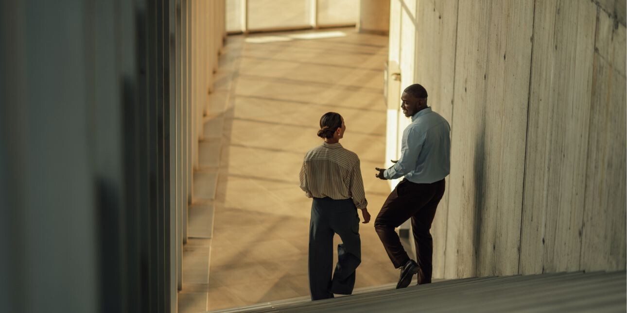 The backs of two business people having a conversation at the bottom of a staircase.