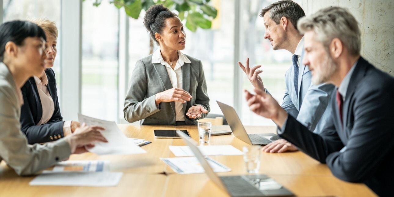 Five board members of different genders and ethnicities engage in a serious discussion around a table in an office.