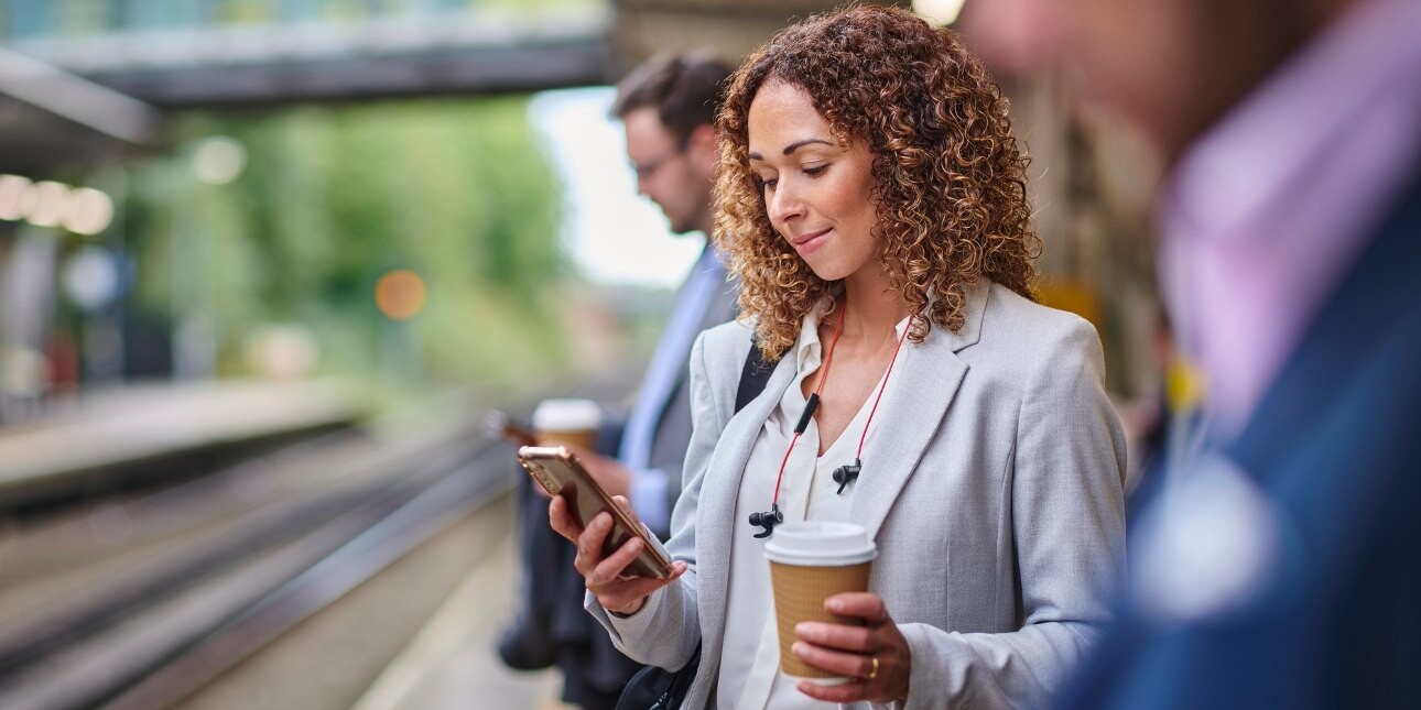 A white woman with brown hair and grey blazer holds a coffee cup in one hand while looking at a mobile in the other hands, while stood on a railway station platform.