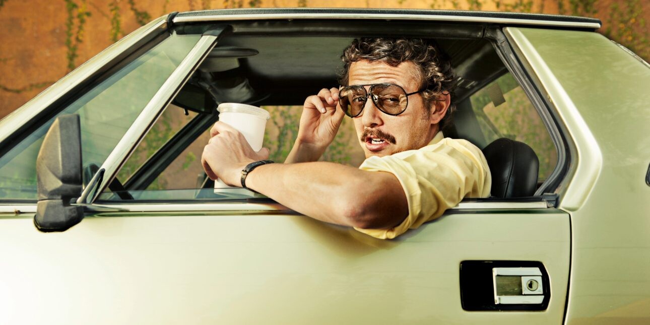 A white man with a moustache and curly hair raises his sunglasses as he leans out of a retro car while holding a coffee cup.