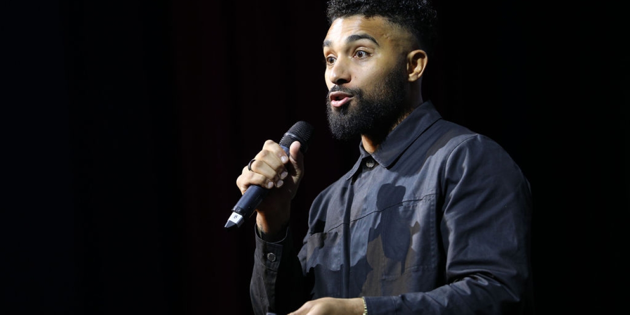 Jay Richards, a black man, wearing a navy jacket and holding a microphone. The background is dark.