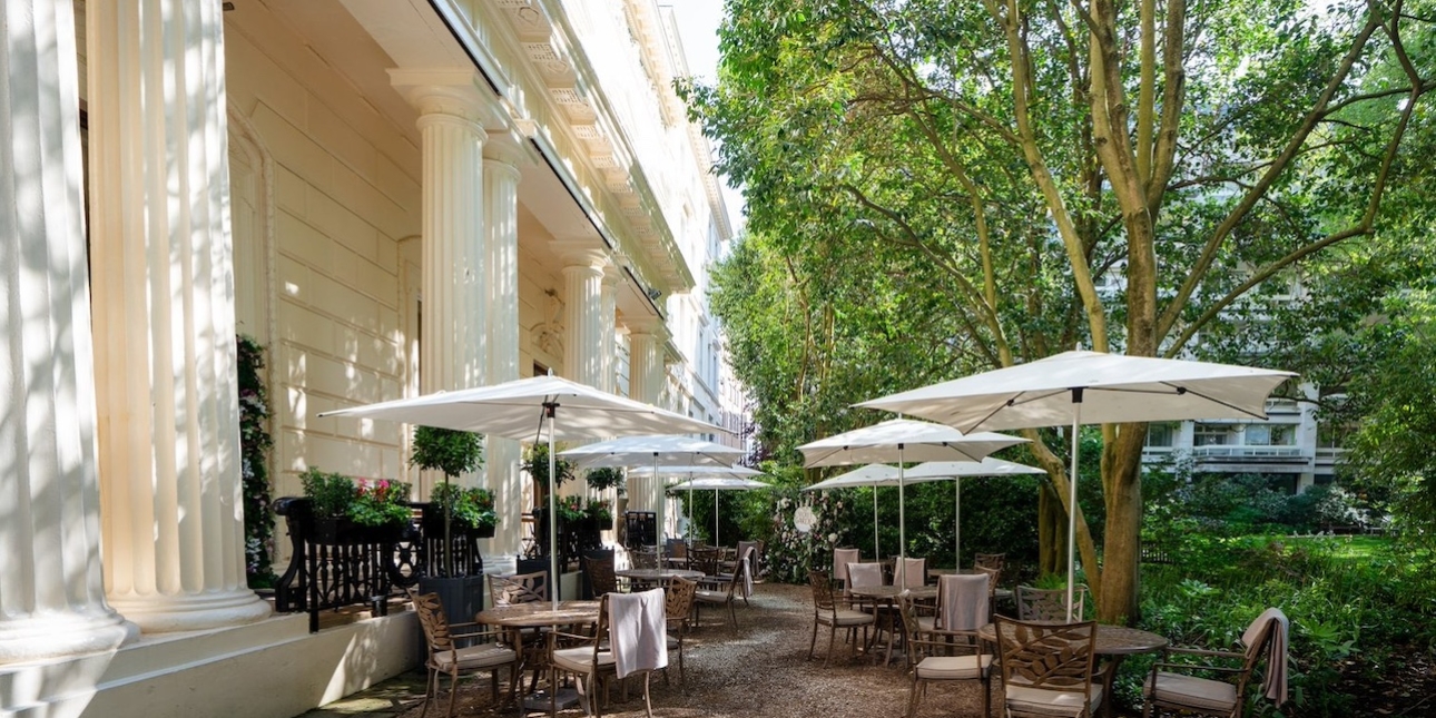 The terrace of 116 Pall Mall consisting of tables and chairs under parasols, leafy green trees on the right and three regency period columns of a cream building
