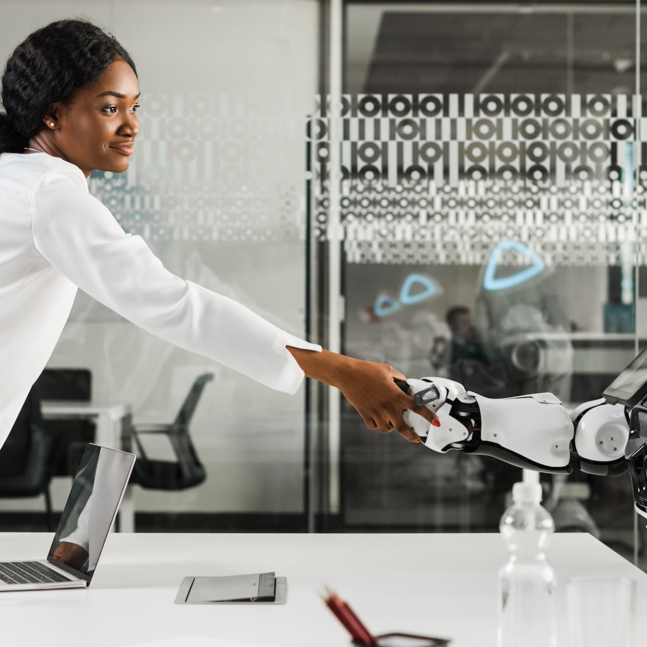In a modern office with glass wall a Black business woman wearing a white shirt and grey trousers shakes with a white humanoid robot