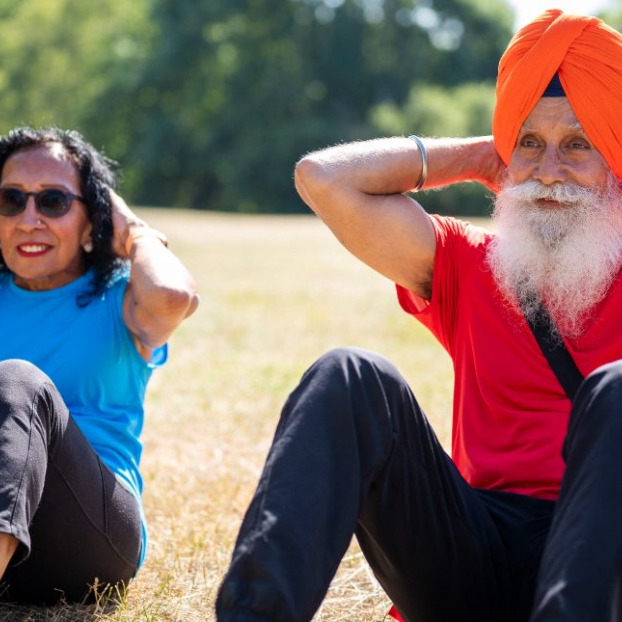 An elderly Asian couple perform sit ups on the grass. On the left is a woman wearing a blue vest and dark leggings. Next to her is a man wearing a red t-shirt, orange vest and blue tracksuit trousers.