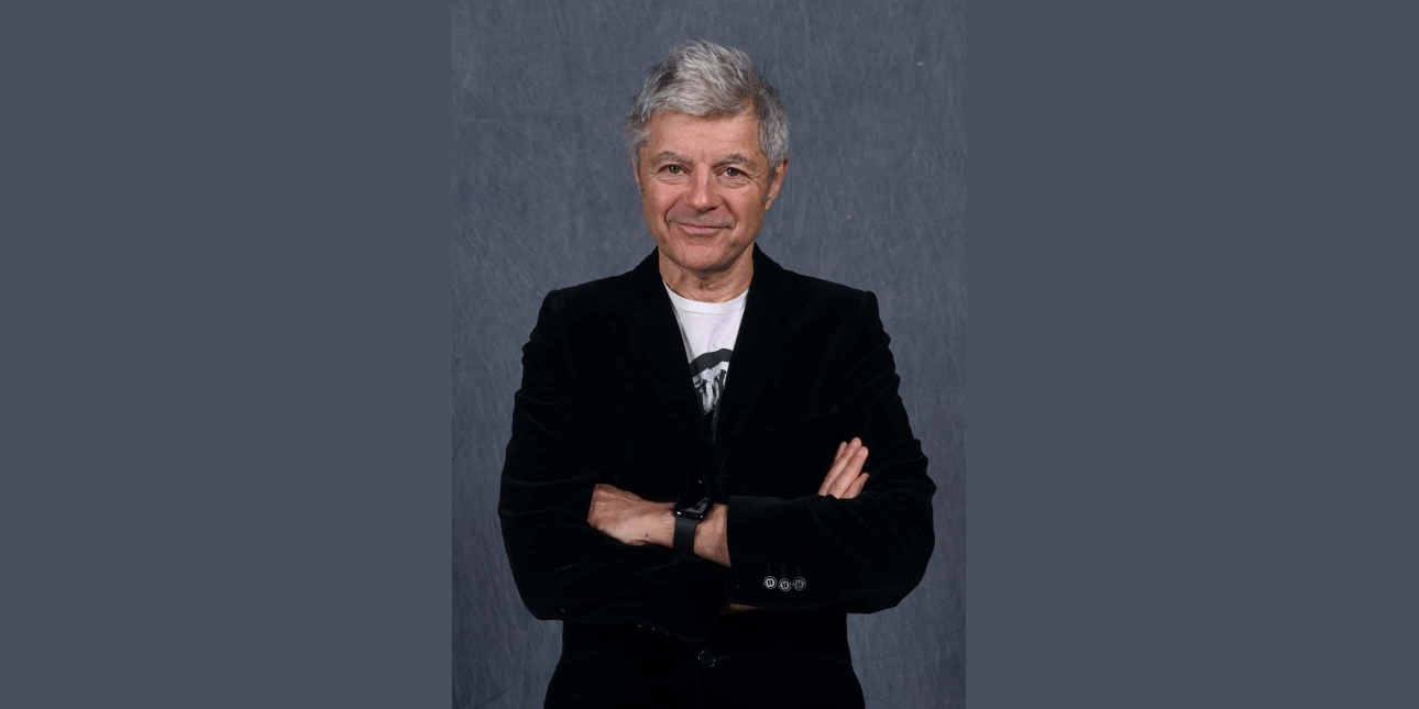 Alan Edwards, a white man with grey hair, wearing a black jacket over a white t-shirt. His arms are crossed. The background is grey