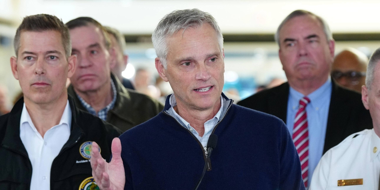 Robert Isom holding a press conference flanked by other officials. Isom is a white middle-aged man with short grey hair andwearing a navy zip-up sweater.