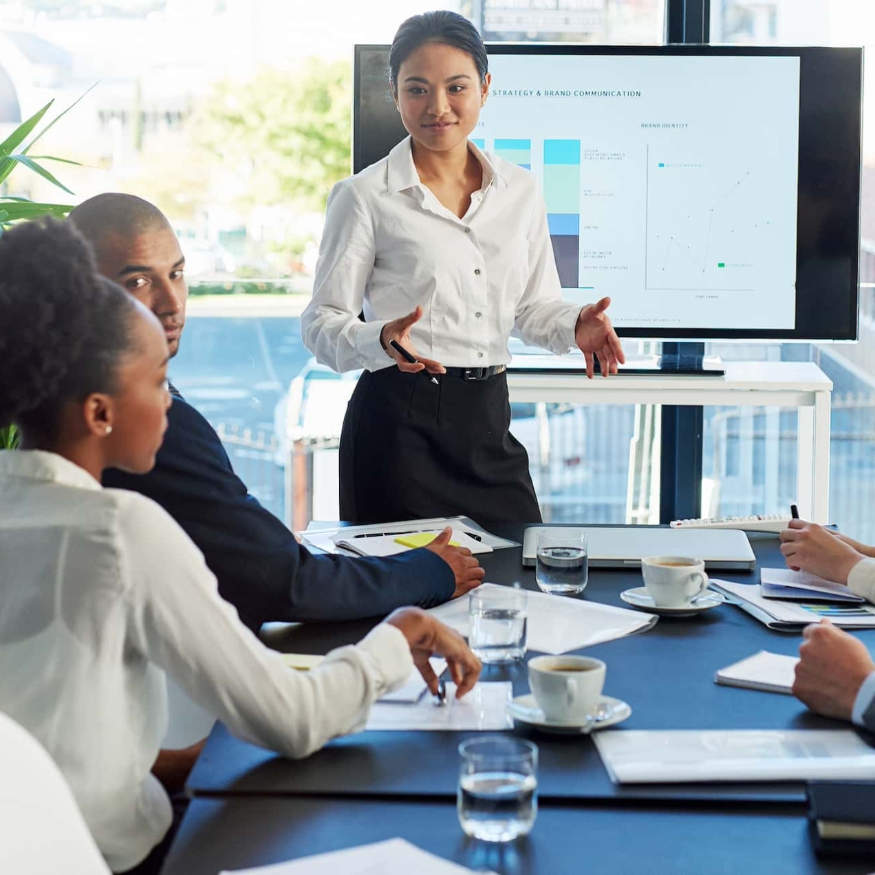 An Asian woman stood in front of a screen addresses fives colleague around the table. They are a Black woman, Black man, white woman and white man. All are smartly dressed