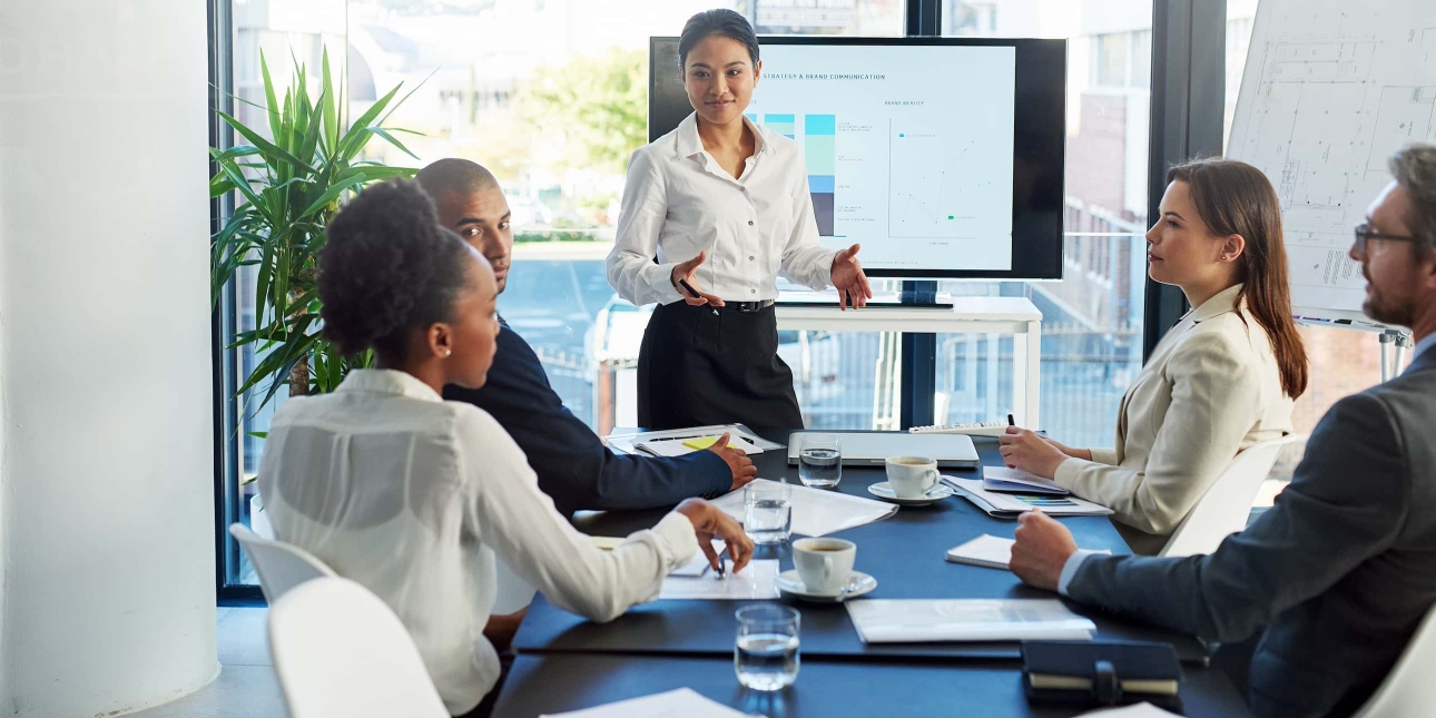 An Asian woman stood in front of a screen addresses fives colleague around the table. They are a Black woman, Black man, white woman and white man. All are smartly dressed