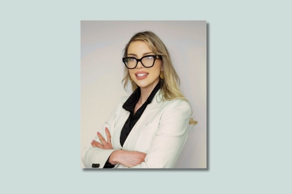 On a grey background, a colour portrait of Amy Simpson. Amy is a white woman with long blond hair who is wearing a light coloured jacket.