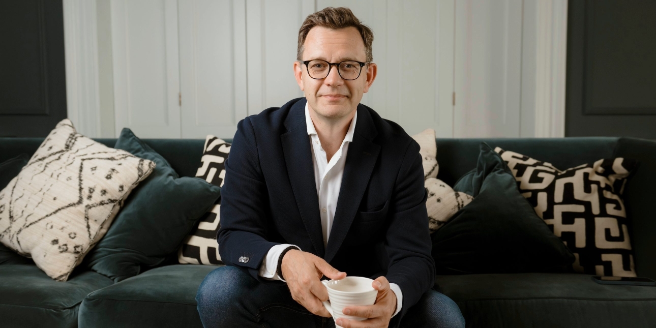 A colour portrait of Andy Coulson sat on a dark sofa with a mug in his hand. Coulson is a white man with short brown hair who wears glasses and a black jacket over a white shirt.