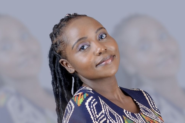 A portrait of Ann-Waithaka on a grey background. Ann is a Black woman with long dark hair who is wearing a navy top with white and grey patterns