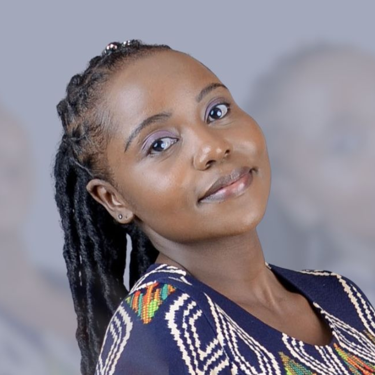 A portrait of Ann-Waithaka on a grey background. Ann is a Black woman with long dark hair who is wearing a navy top with white and grey patterns