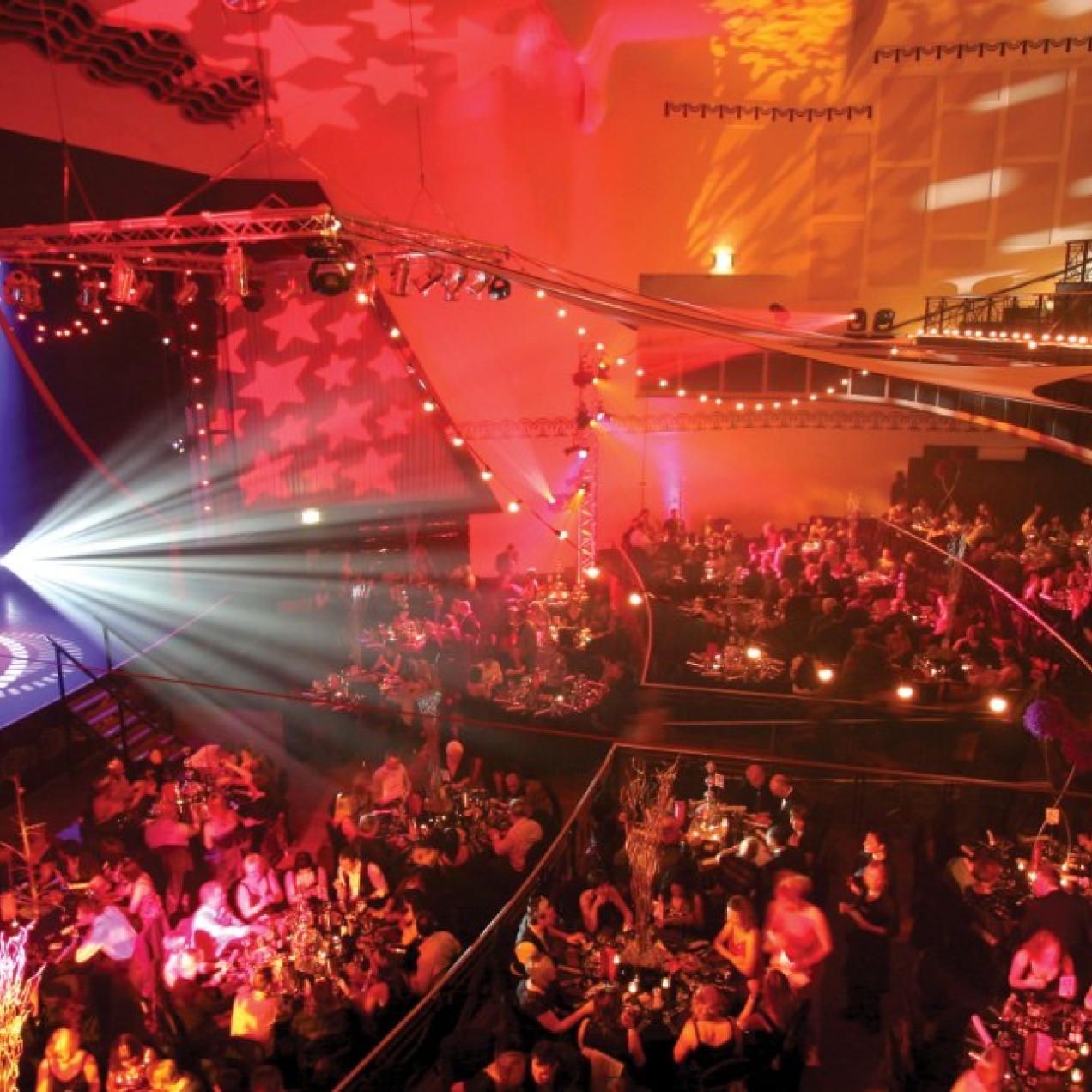 A colourful photo taken from a balcony overlooking the large auditorium and stages were audiences are sat around dozens of tables.