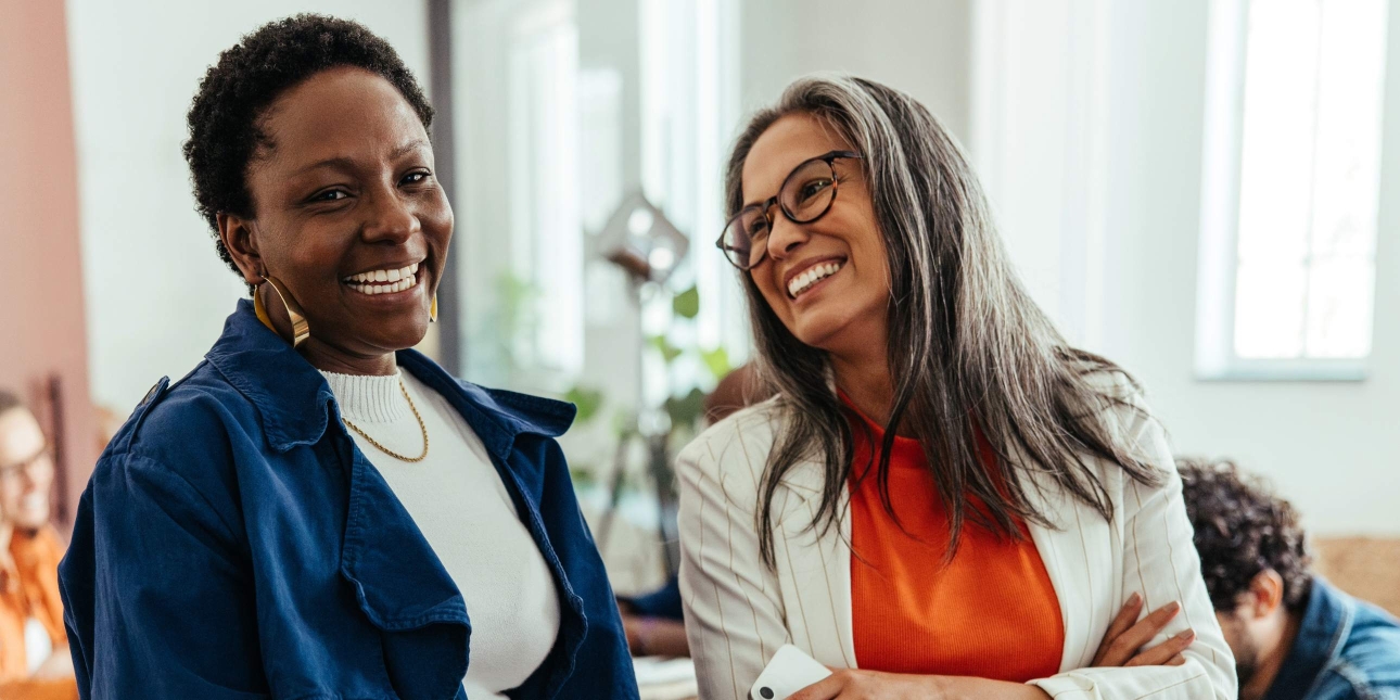 Two women in an office smiling mid-conversation. On the left is a Black woman with short dark hair wearing a blue jacket over a white top. On the right is an older white woman with long greying brown hair and glasses wearing a cream jacket over an or