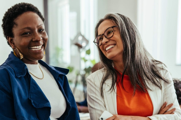 Two women in an office smiling mid-conversation. On the left is a Black woman with short dark hair wearing a blue jacket over a white top. On the right is an older white woman with long greying brown hair and glasses wearing a cream jacket over an or