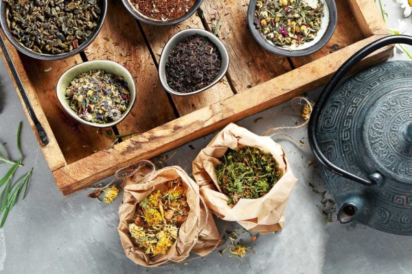 A bird's eye view of a grey teapot and various types of teas in small bowls on a grey stone surface.