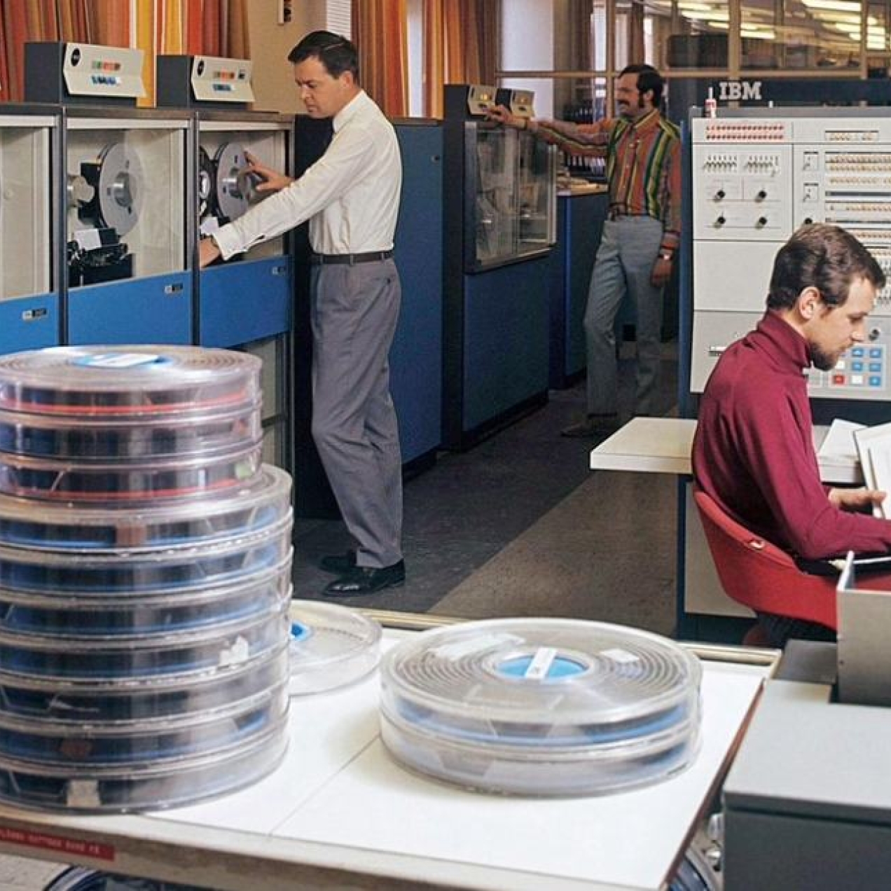 A photograph from 1966 showing three white men inside a room with early and large IBM computing equipment, stacks of data-tape reels and computer terminals.