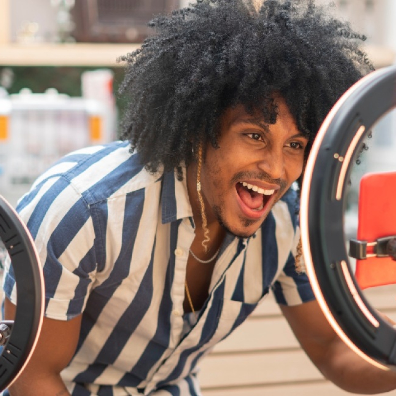 A smiling young Black man wearing blue and white striped shirt while crouching down in front of a mobile phone on a tripod which is flanked by two ring flashes.