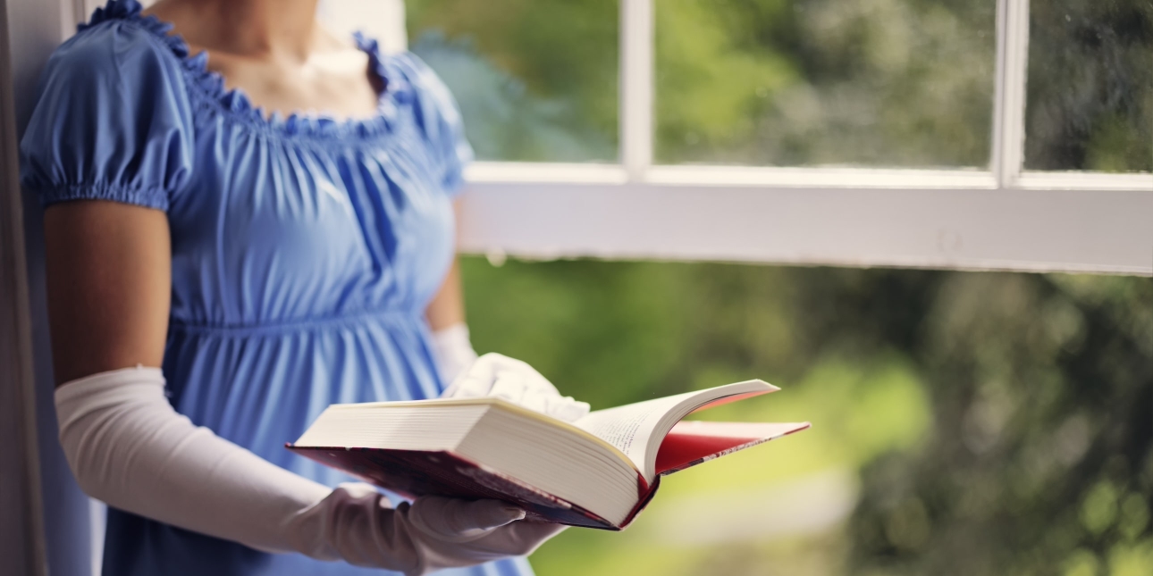 A cropped shot of an unidentified woman wearing a Regency style dress and white gloves, stood reading a book beside a large open sash window