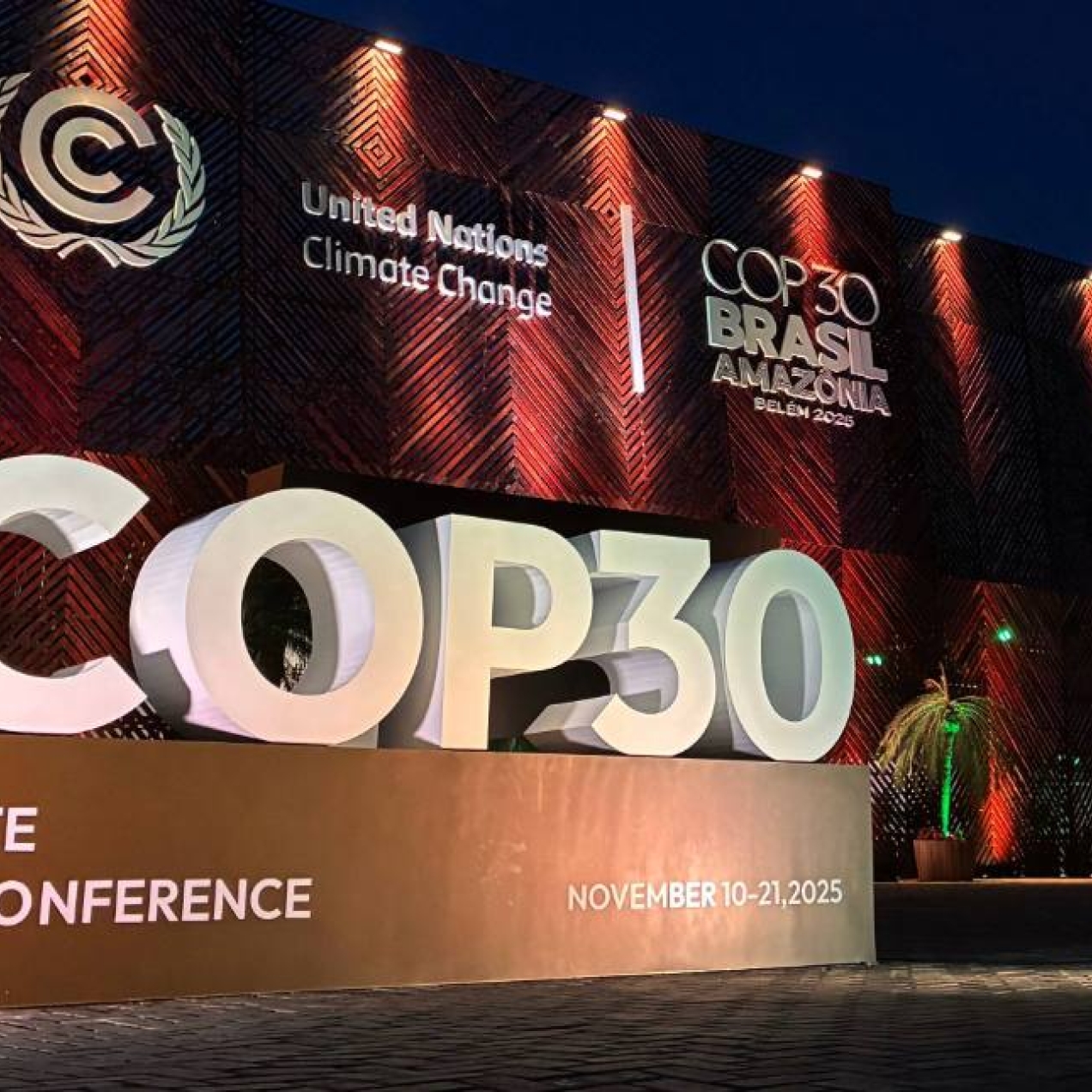 Lit up against a dark night's sky, a three-dimensional typographic sculpture featuring a large hashtag COP30 on a plinth in front of the main entrance venue in Belém, Brazil. Various logos feature in white.