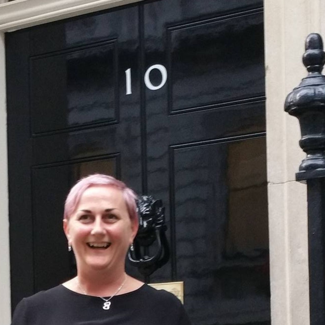 Carolyne Mitchell stood on the steps of 10 Downing Street. Carolyne is a white woman with short blonde hair wearing a black top. The door is back with white numbers