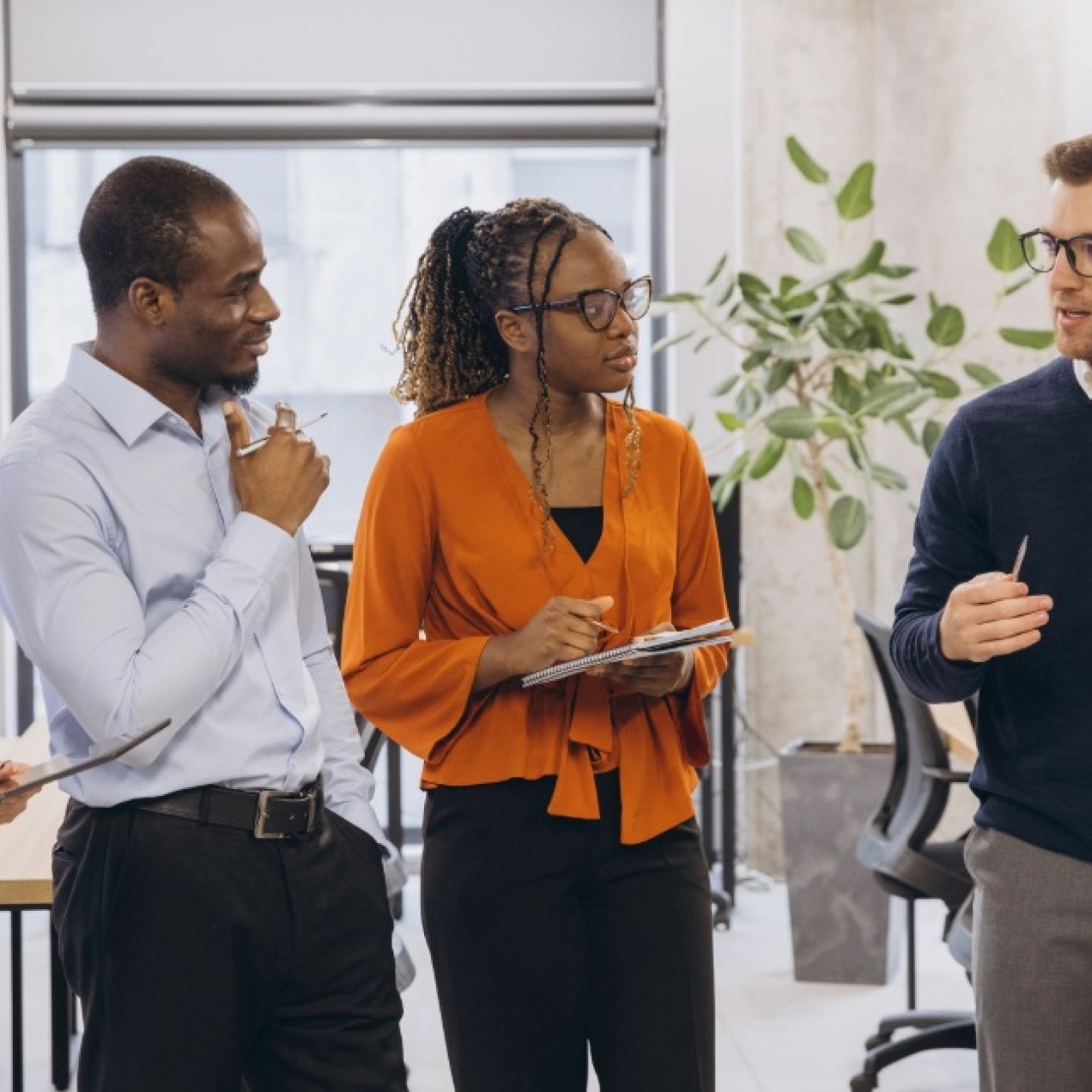 Four colleagues in smart-casual clothing stood in a modern office discussing sticky notes on a cement wall. From left to right: a white woman, a Black man, a Black woman, and a white man.