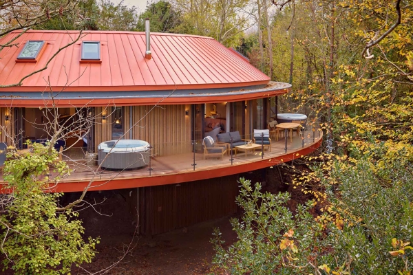 Exterior of a semi circular accommodation with red roof, wooden walls, balcony containing loungers and plunge pools, on stilts surrounded by tall leafy trees