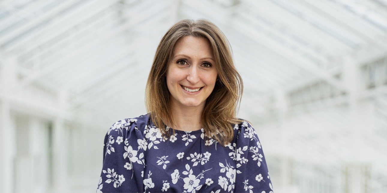 A portrait of Christine Richardson against the backdrop of a white pitched ceiling. Christine is a white woman with long dark hair who wears a blue top decorated with white florals
