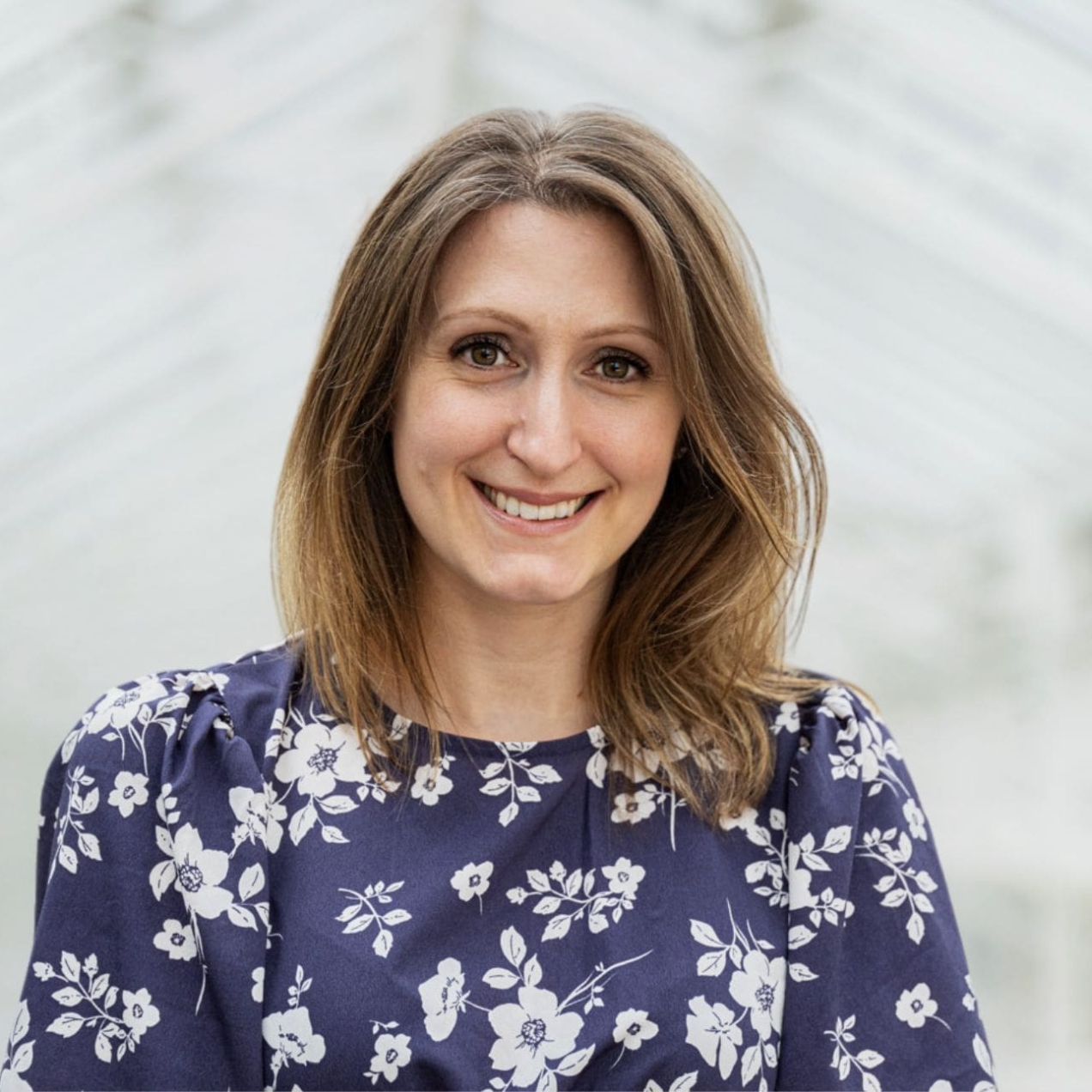 A portrait of Christine Richardson against the backdrop of a white pitched ceiling. Christine is a white woman with long dark hair who wears a blue top decorated with white florals