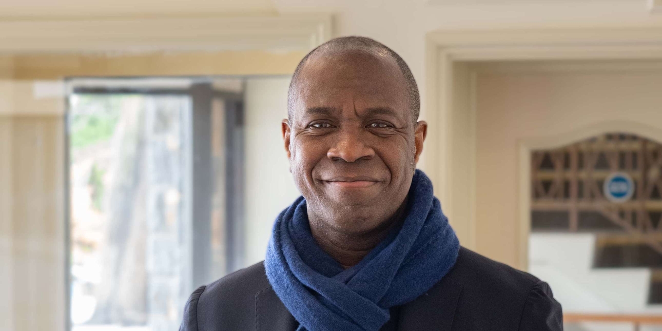 A portrait of Clive Myrie. Clive is a Black man who looks directly at the camera smiling. He wears a navy scarf. The background is a magnolia room.