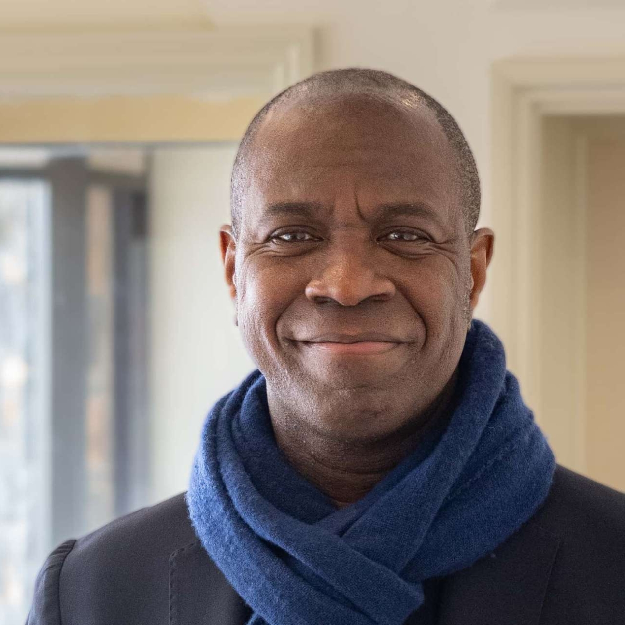 A portrait of Clive Myrie. Clive is a Black man who looks directly at the camera smiling. He wears a navy scarf. The background is a magnolia room.