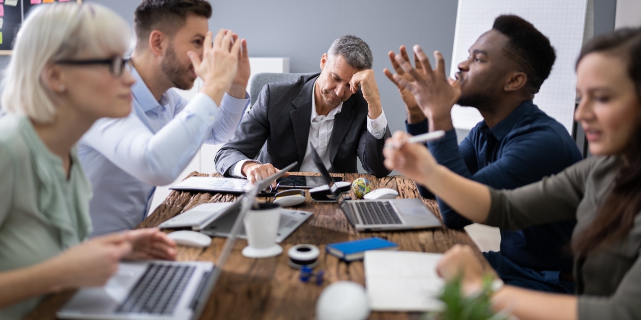 A suited white man leans his elbow on the table and rests his head while four colleagues of different genders, ages and ethnicities wave their arms in an angry discussion