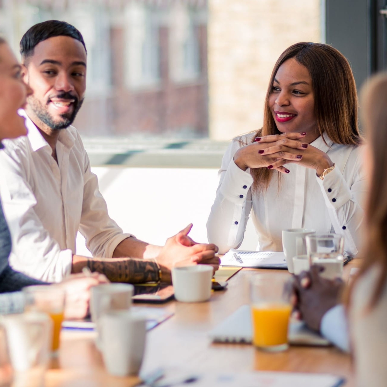 Six colleagues around board table. In focus are a white woman with blonde hair and blue shirt, a Black man with short dark hair and cream shirt, and a Black woman with brown hair and white shirt