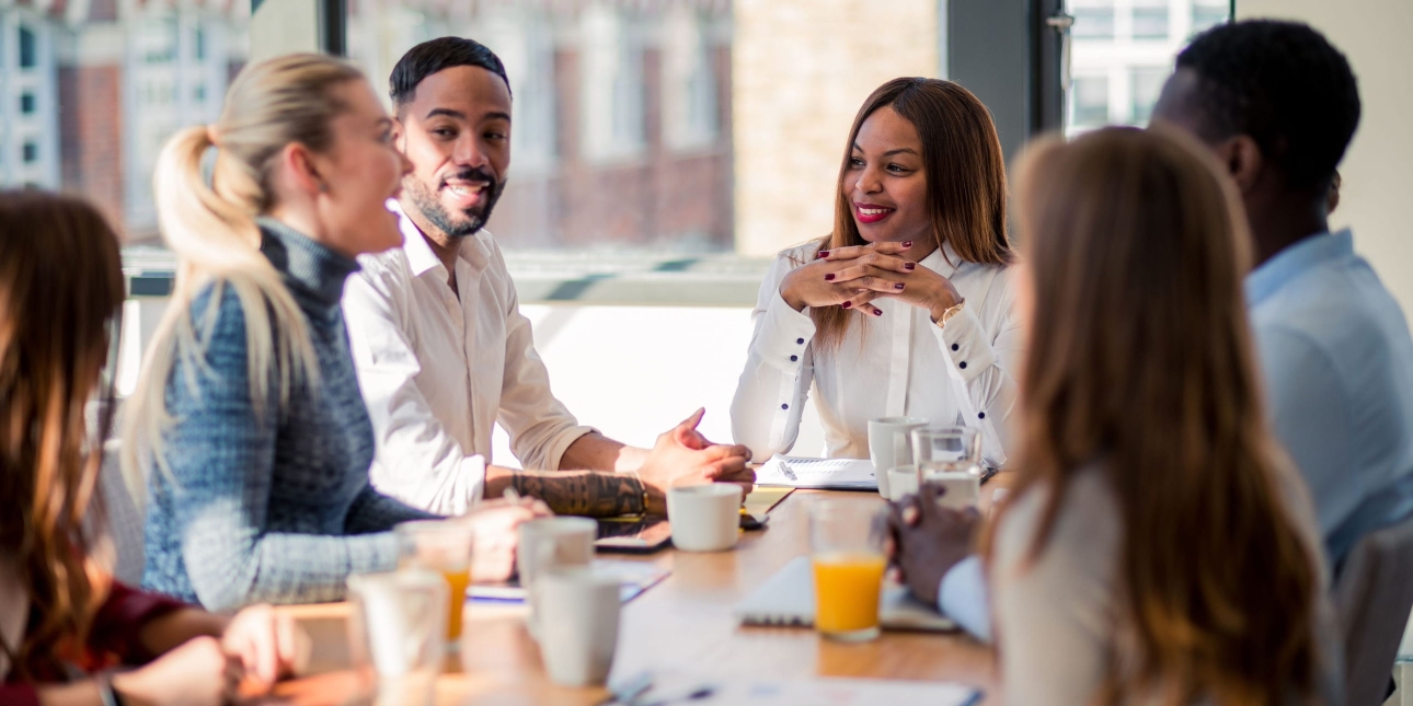 Six colleagues around board table. In focus are a white woman with blonde hair and blue shirt, a Black man with short dark hair and cream shirt, and a Black woman with brown hair and white shirt