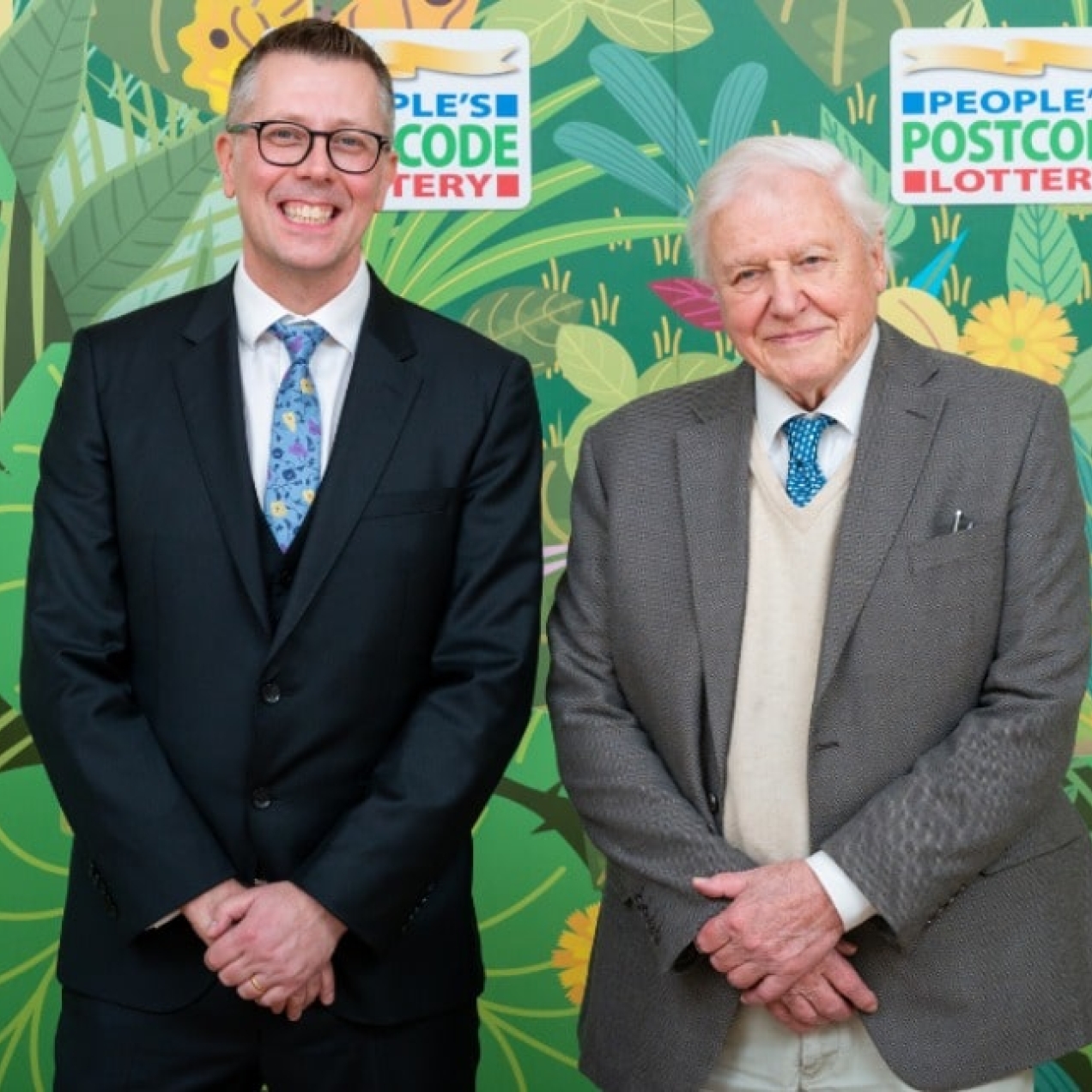 David Miller and Sir David Attenborough stood against a background composed of illustrated leaves and fauna. Both are white men with short hair wearing suits.